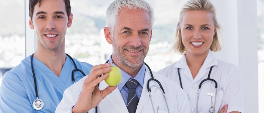 Krankenversicherung - zwei junge Ärzte stehen neben einem Oberarzt, der einen Apfel in der Hand hält.